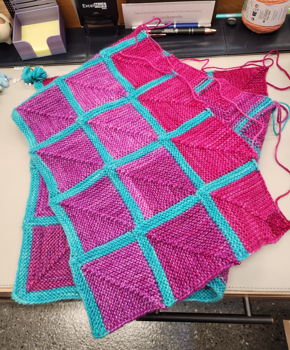 A blanket in progress made of many pink and aqua blue squares, folded to fit on a desk.