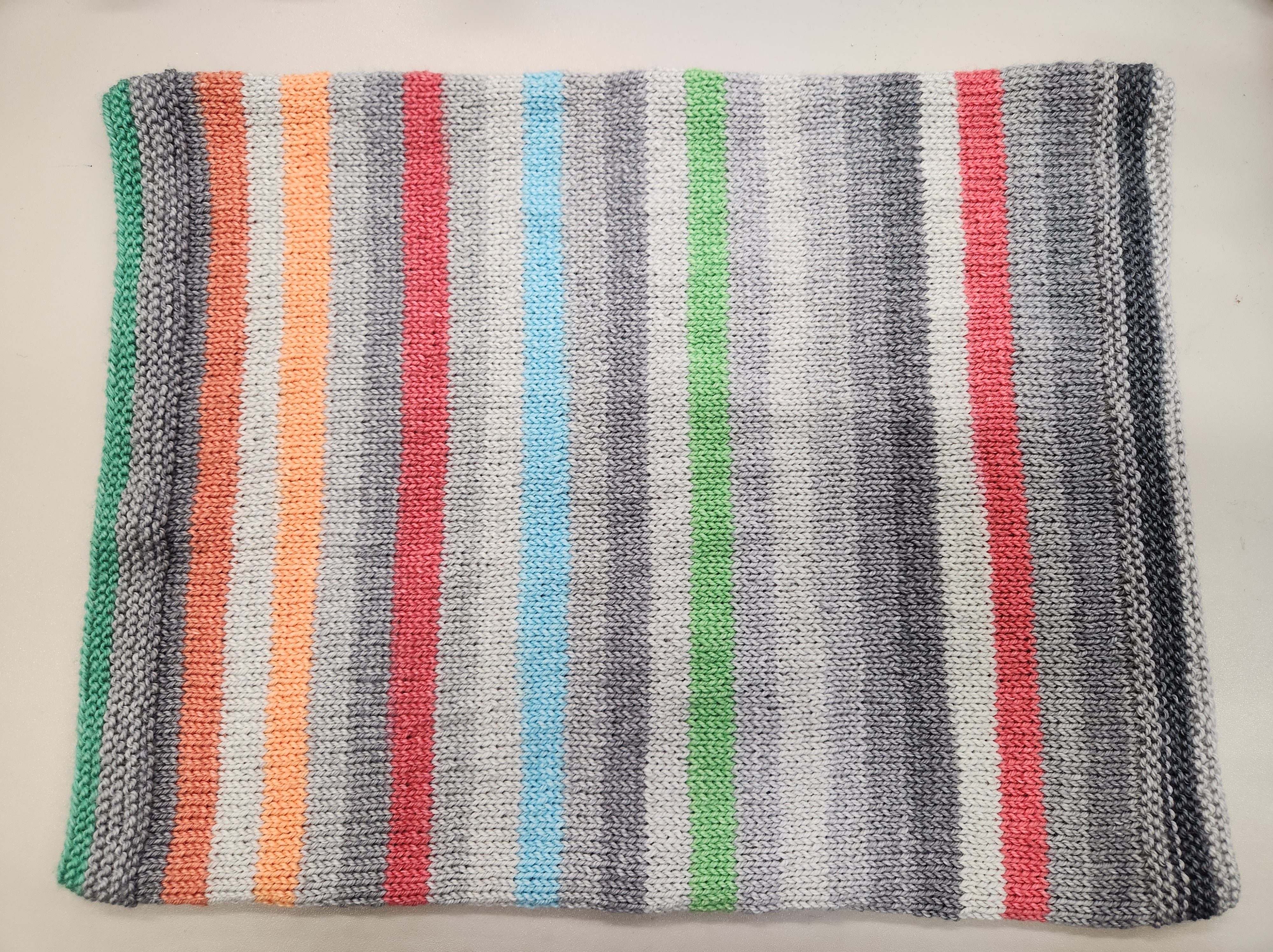 A knitted cowl in stripes of green, grey, orange, red, and blue on a grey background.