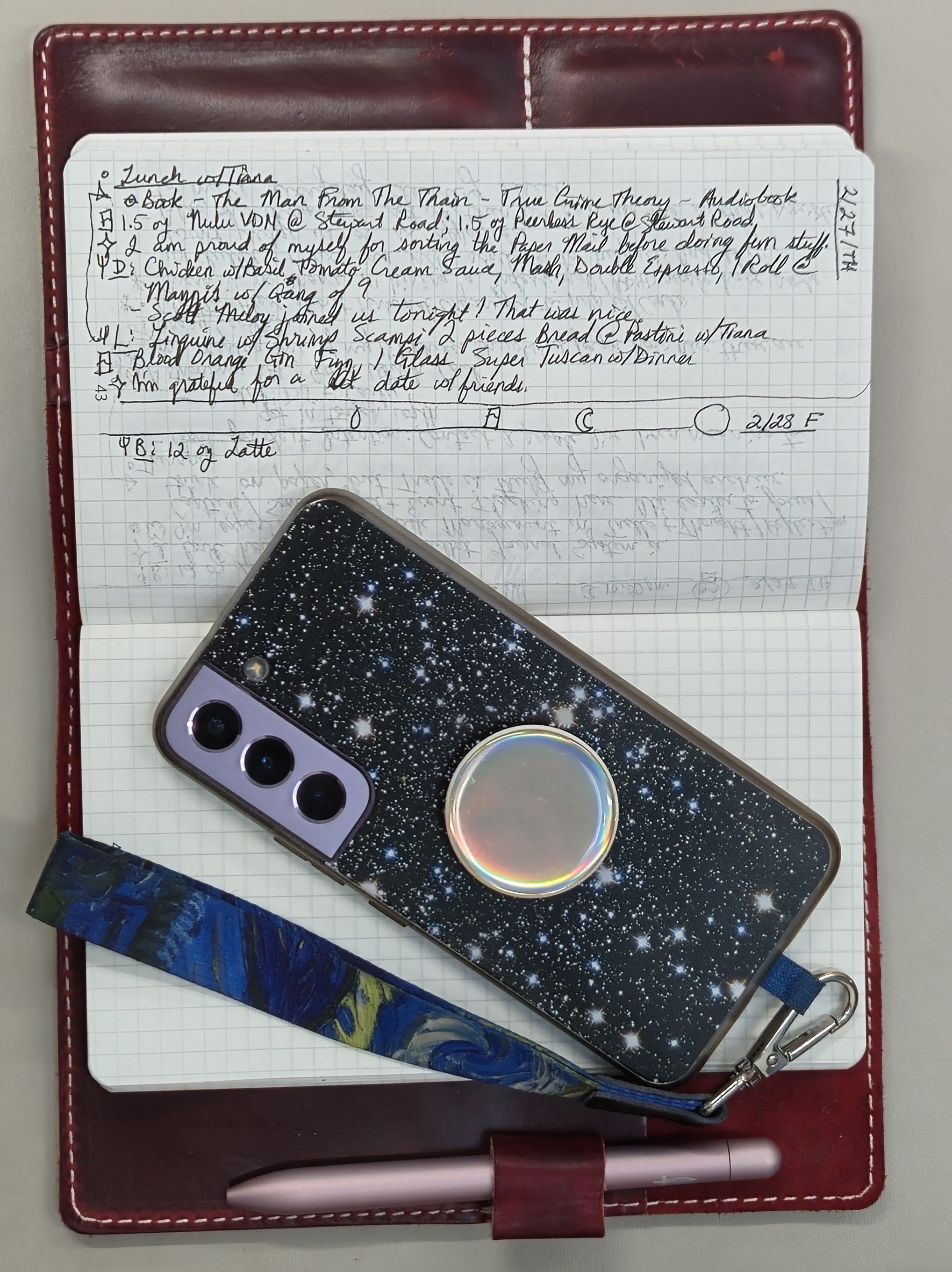 A grid-ruled notebook in a leather cover lies open. You can see some notes in black pen. A phone in a star-patterned case with an iridescent Pop Socket on the back and a wrist loop lies on top of the notebook.